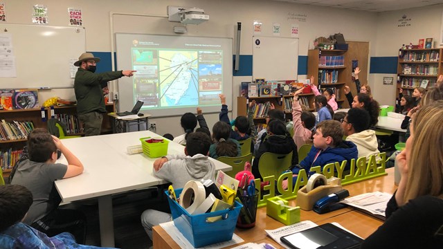 Park ranger pointing as 4th-grade students raise hands in a classroom, a powerpoint on a screen