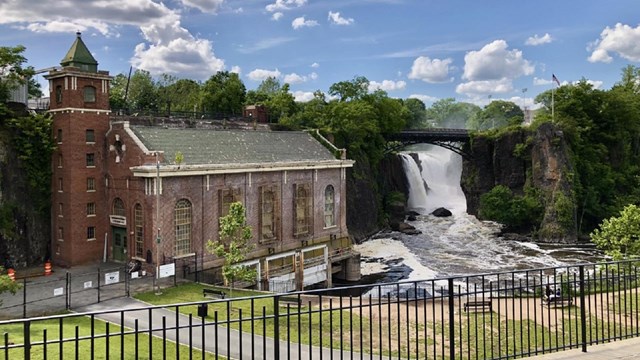 A 77 ft. waterfall framed by an arched bridge sits beside a brick hydroelectric power plant