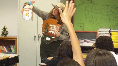 Park Ranger in a classroom showing students the NPS symbol.  A student's raised hand in foreground.