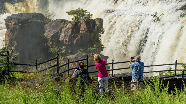 Visitors hold phones & cameras up at a roaring waterfall overlook