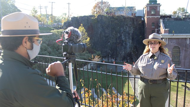 A park ranger smiles as she gives a presentation outside while another ranger films her