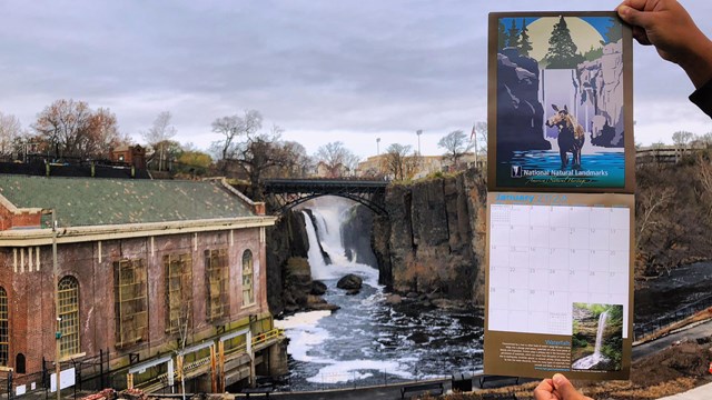 Hands hold a National Natural Landmarks January calendar page of a waterfall before the Great Falls