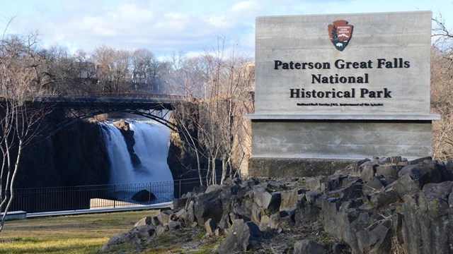 Rectangular concrete sign for Paterson Great Falls NHP stands before a waterfall framed by a bridge