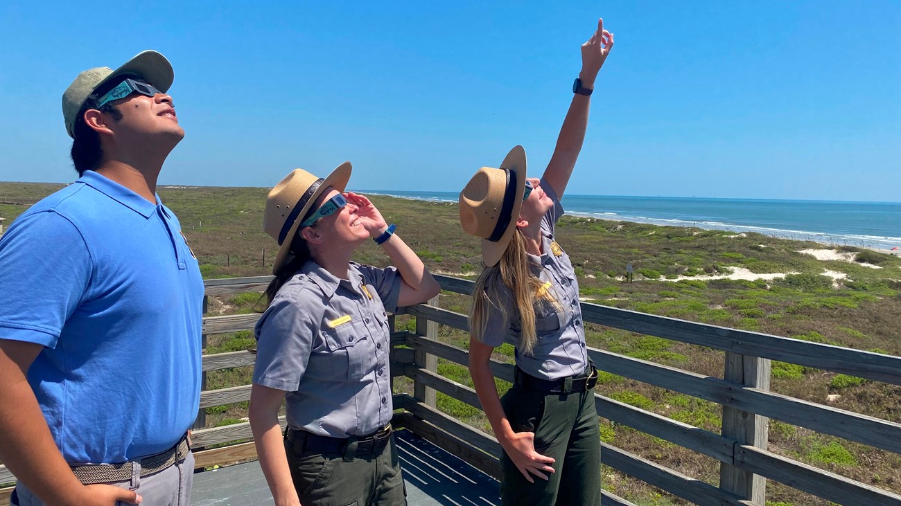 Three people wearing eclipse glasses look up at the sun. 