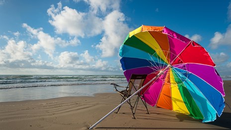 A rainbow umbrella lies open on the beach next to a chair with the waves and blue sky in front.