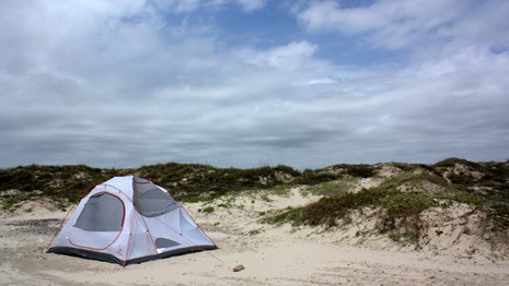 White tent on the beach with vegetated dunes behind and a cloudy-blue sky above.