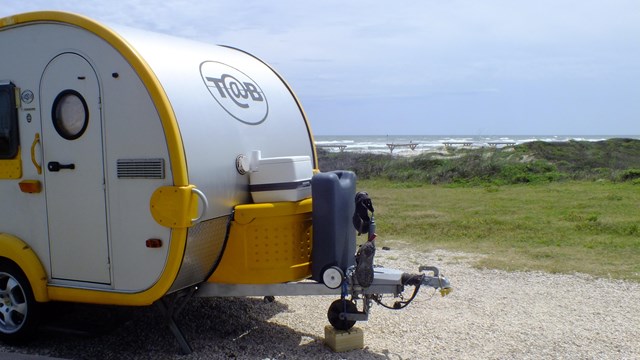 Silver and yellow camping trailer on the left with the beach visible in the distance.