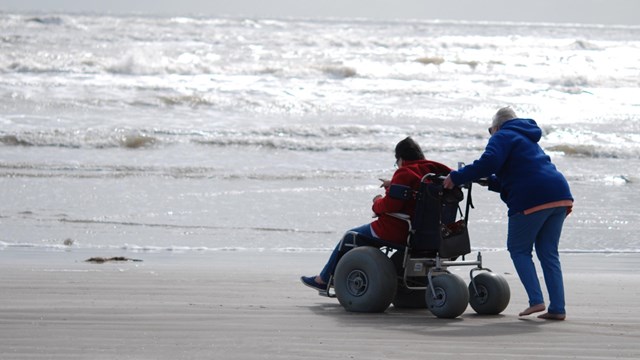 A person pushes another person sitting in a beach wheelchair. 