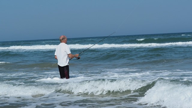 A man standing in the shallow ocean water while fishing. 