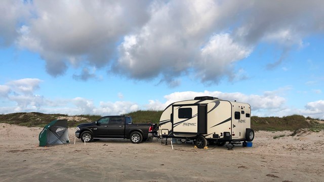 A truck with trailer is parked on the beach with the dunes behind.