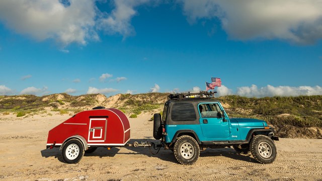 A blue jeep pulling small, red trailer is parked on the beach with the dunes behind.