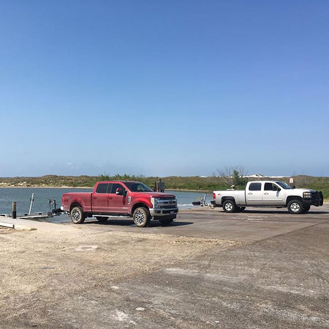 Two trucks with trailers backed up to calm water.  There is a small dock in the water.