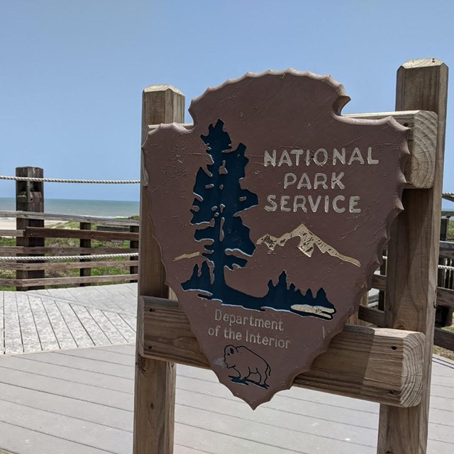 National Park Service Arrowhead on wooden posts with the beach and gulf available in the distance.