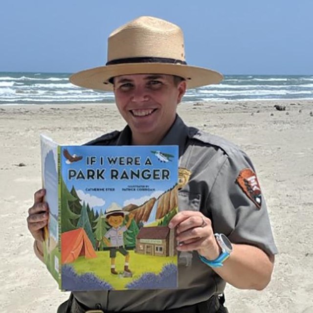 A ranger stands on the beach with the gulf behind holding a book titled 