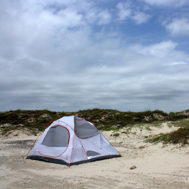 White tent on the beach with vegetated dunes behind and a cloudy-blue sky above.