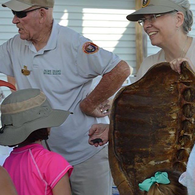 Two volunteers stand in front of a group of people, one volunteer is holding a sea turtle shell.