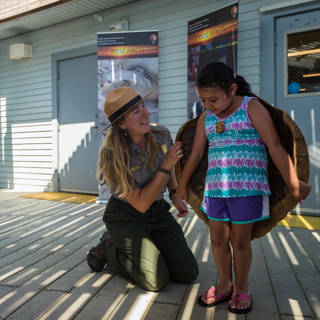 A ranger kneels next to a girl helping her hold up a turtle shell on her back.