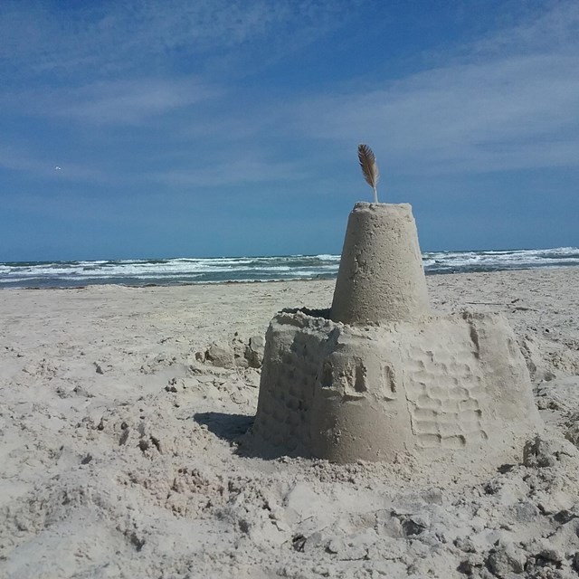 A sand castle it a feather in the top is on the right in the foreground, with the ocean in the back.