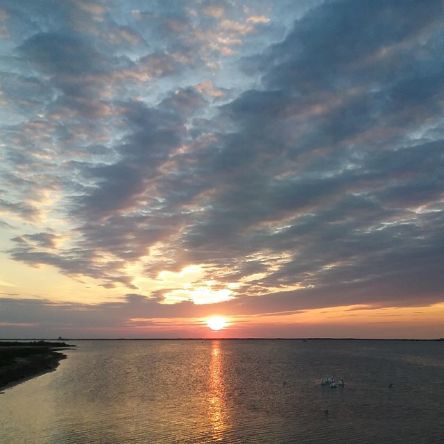 A sunset over a body of water with streaky clouds and the orange sun just barely visible below.