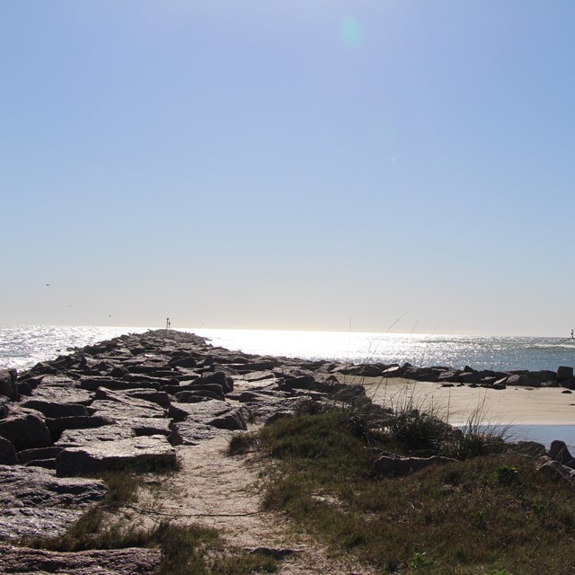 A rock jetty is on the left side with water on both sides of it.  It is a sunny day.