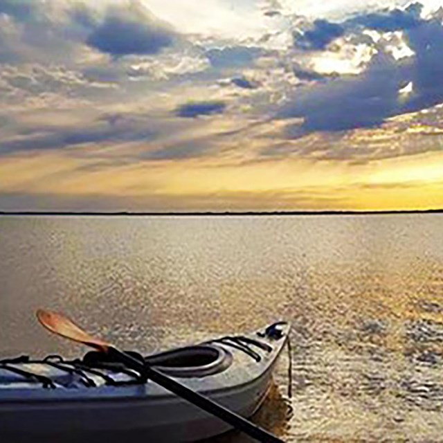 A kayak rests partly on shore on the left in calm water with a blue and yellow sunset sky above.