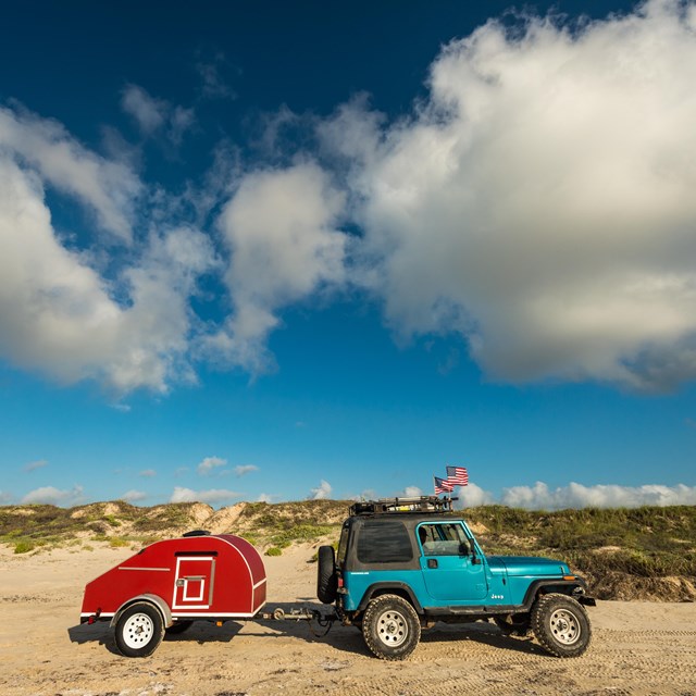 A blue jeep pulling small, red trailer is parked on the beach with the dunes behind.