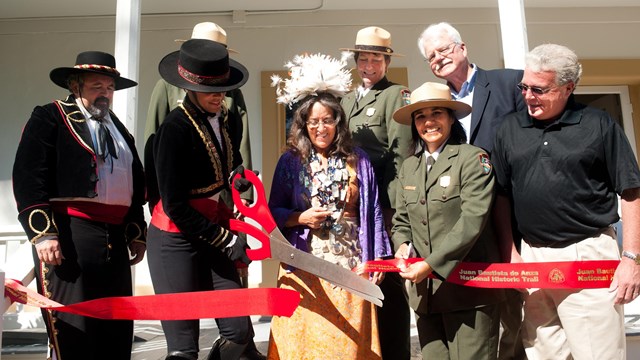 Two people wearing Spanish colonial clothing cut a red ribbon with large scissors.
