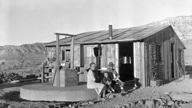 Black and white photo of a family sitting on a well in front of a small wooden ranchhouse.