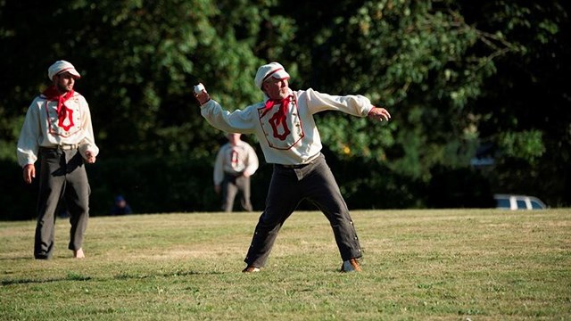 Man dressed in white shirt with red scarf and white hat holds a small white ball above his head.