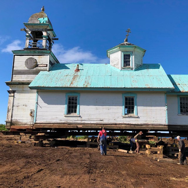 A white church with green roof up on wooden casters next to cliff overlooking ocean