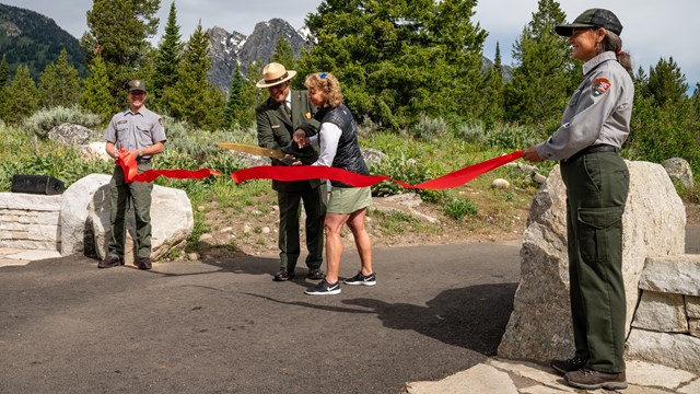 NPS Rangers hold red ribbon aloft against backdrop of mountains & trees. Ribbon is being cut. 