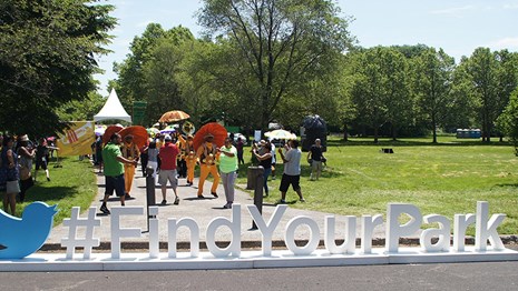 Large Find Your Park hashtag sign in a city park with people dancing behind it with umbrellas.