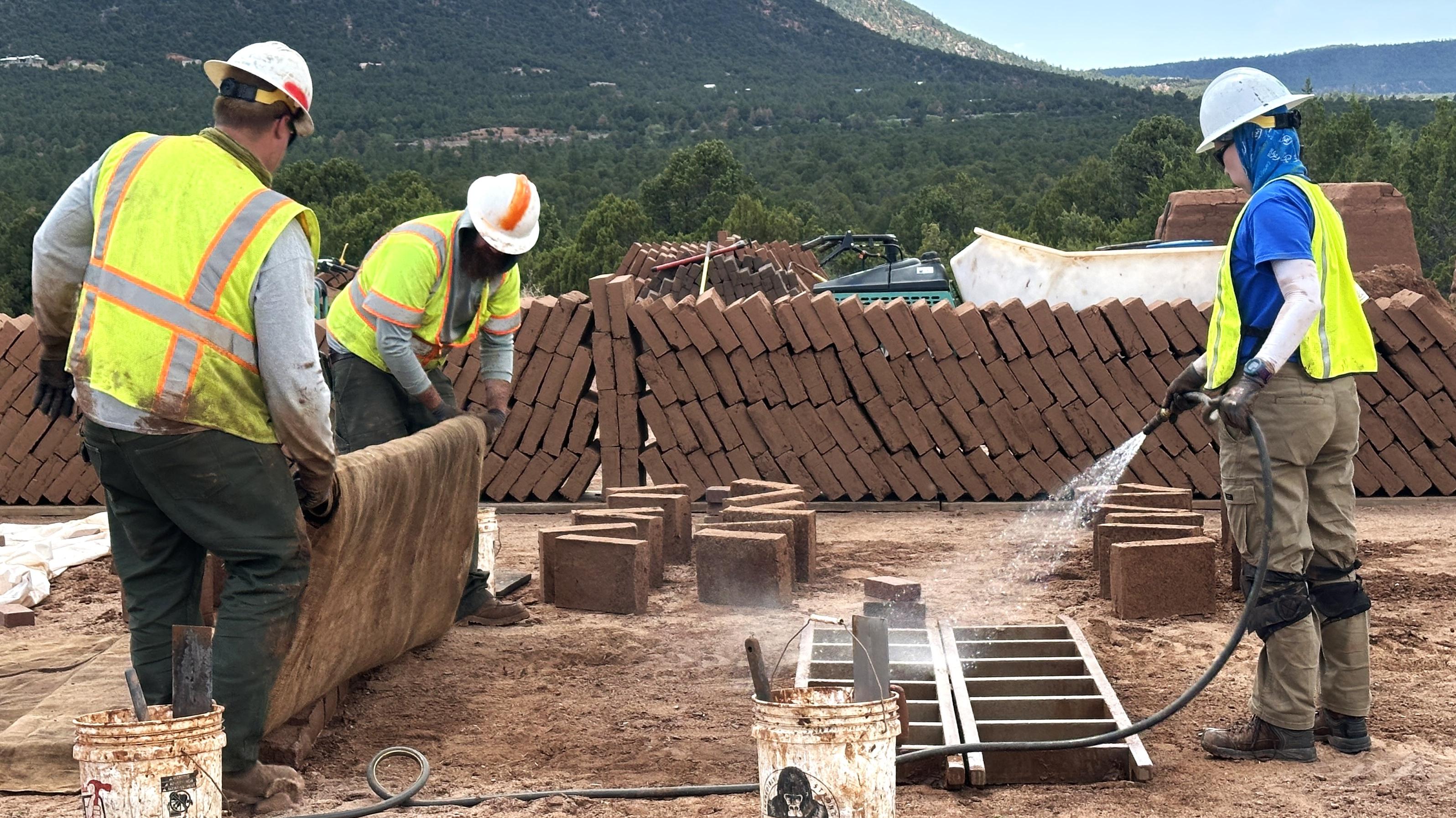 Two workers in yellow vests hold fabric next to a worker spraying adobe mold, brown bricks behind