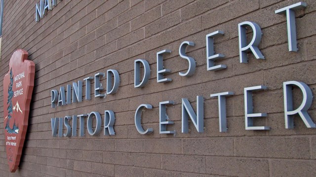 the side of a tan building with the park service logo and text: "painted desert visitor center."