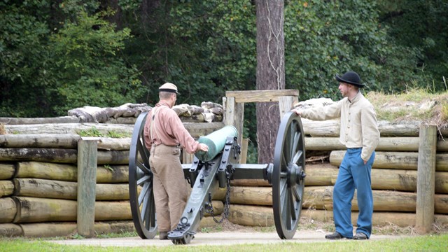 Two white men wearing Confederate uniforms stand next to a cannon in front of a wooden half wall.