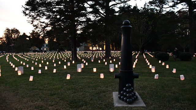 The glow of luminaries front of headstones is just visible under an orange and yellow sunset.