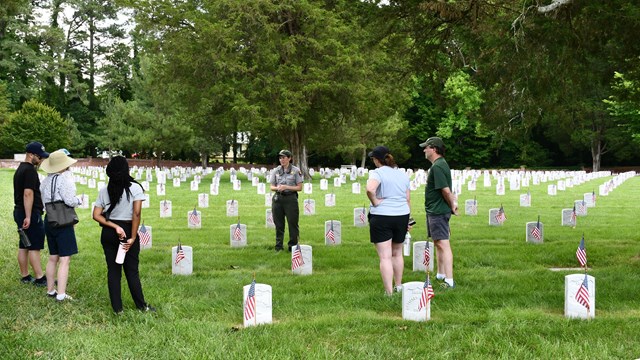White woman in NPS uniform speaks to a group in a cemetery with flags in front of the headstones.