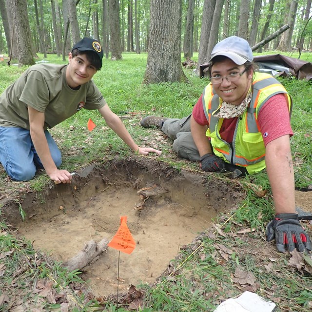 Two young interns excavate a square hole, smiling at camera.