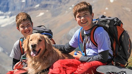 Two kids wearing backpacks sit with a leashed golden retriever dog