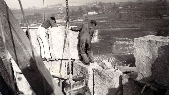 a black and white photo of 2 men standing on stacked blocks of granite while receiving another block