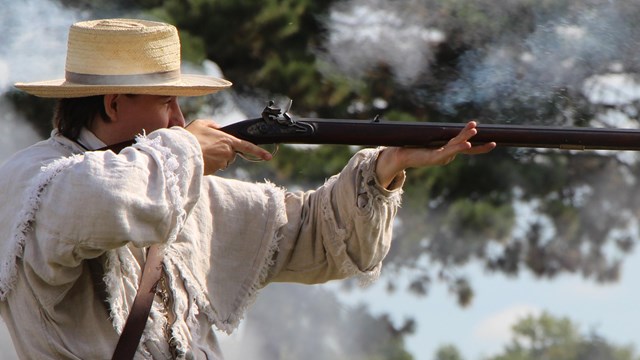 man wearing a straw hat and white overjacket shooting a rifle from era 1812