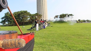 Line of people in War of 1812 fire muskets. In the foreground is a muzzle of a carronade.