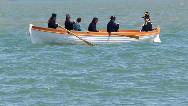 six people in 1812 costumes sit in a wooden boat with oars at rest