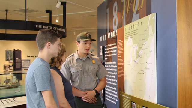 A woman in a blue shirt listens to a park ranger who points to a wall map of the War of 1812