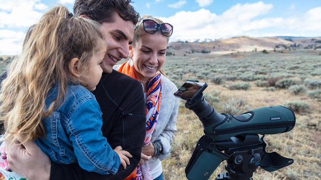 A man, woman and child look into a phone mounted to a spotting scope in a sagebrush covered valley