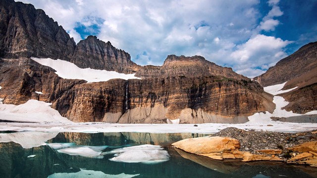 Photo of snow partially melted on a lake surrounded by dramatic rock formations.