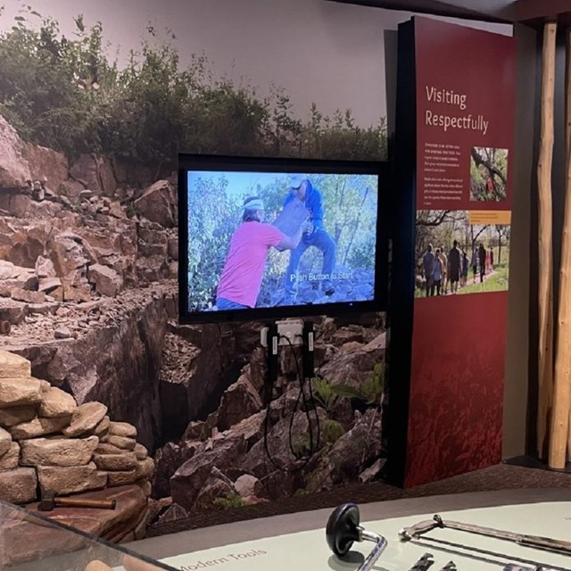 An exibit with stones and tools on a table with a tv screen in the background amid displays