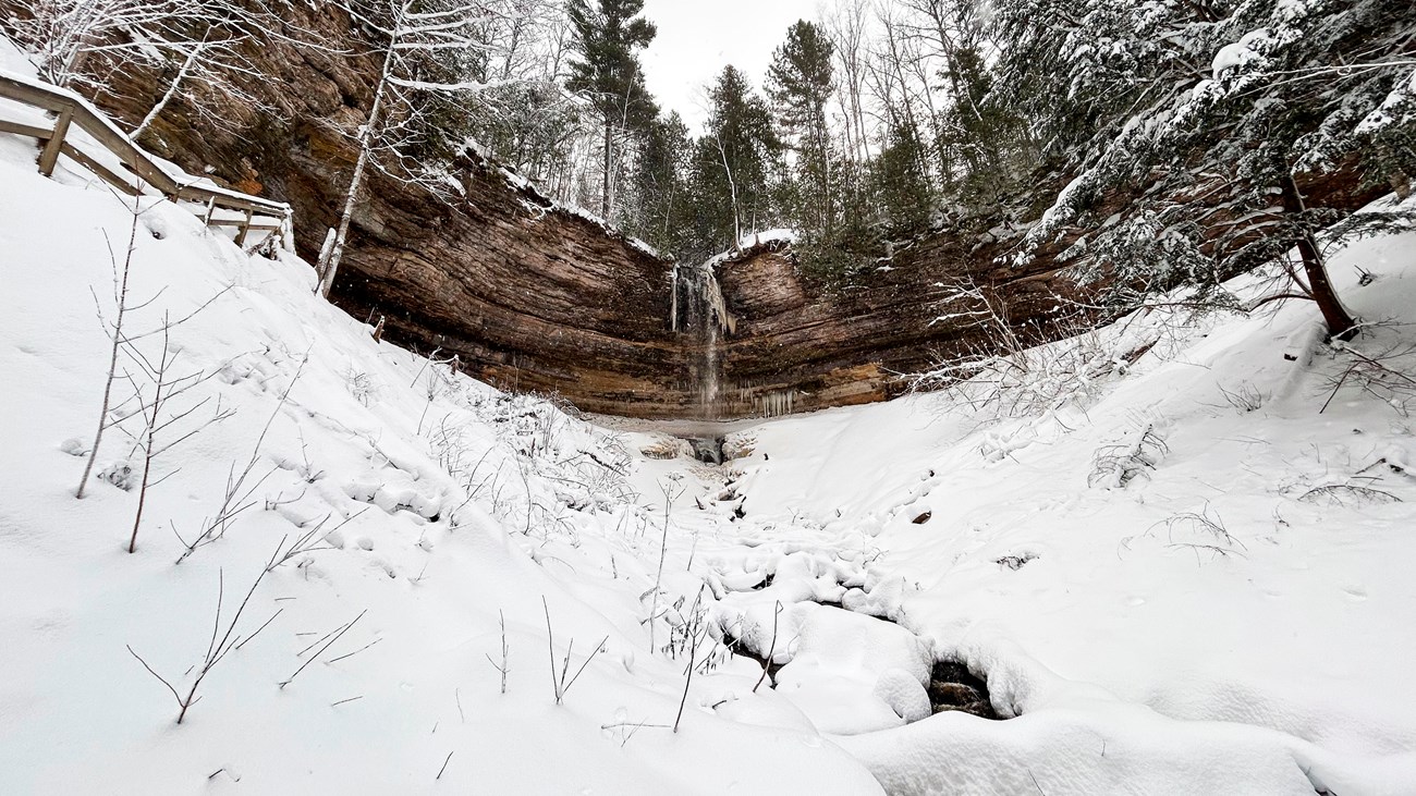 a partially frozen waterfall cascades over a sandstone cliff surrounded by a thick layer of snow.