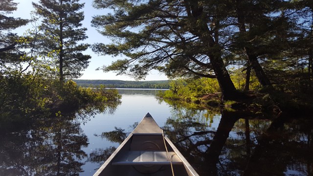 The front of a canoe is seen from the perspective of the viewer facing trees and a lake.