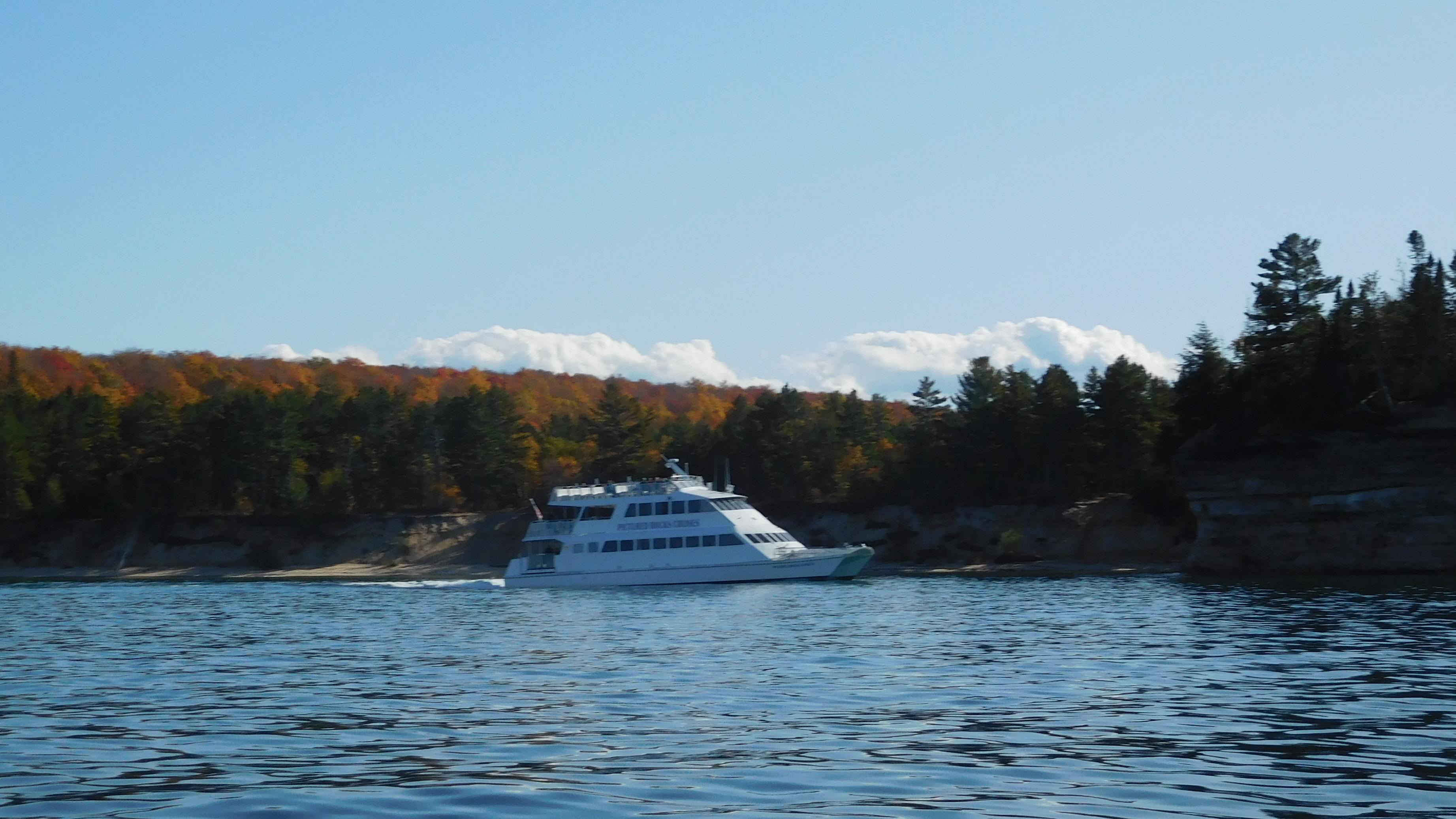A white cruise ship sits in the water in front of autumn foliage.